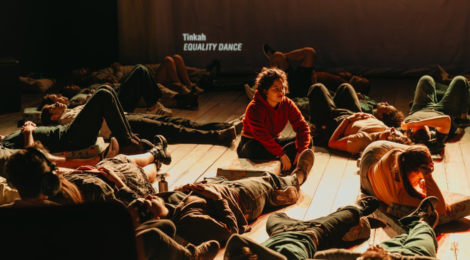 Participants lie and sit across a dimly lit stage during an immersive performance, illuminated by warm amber lighting.